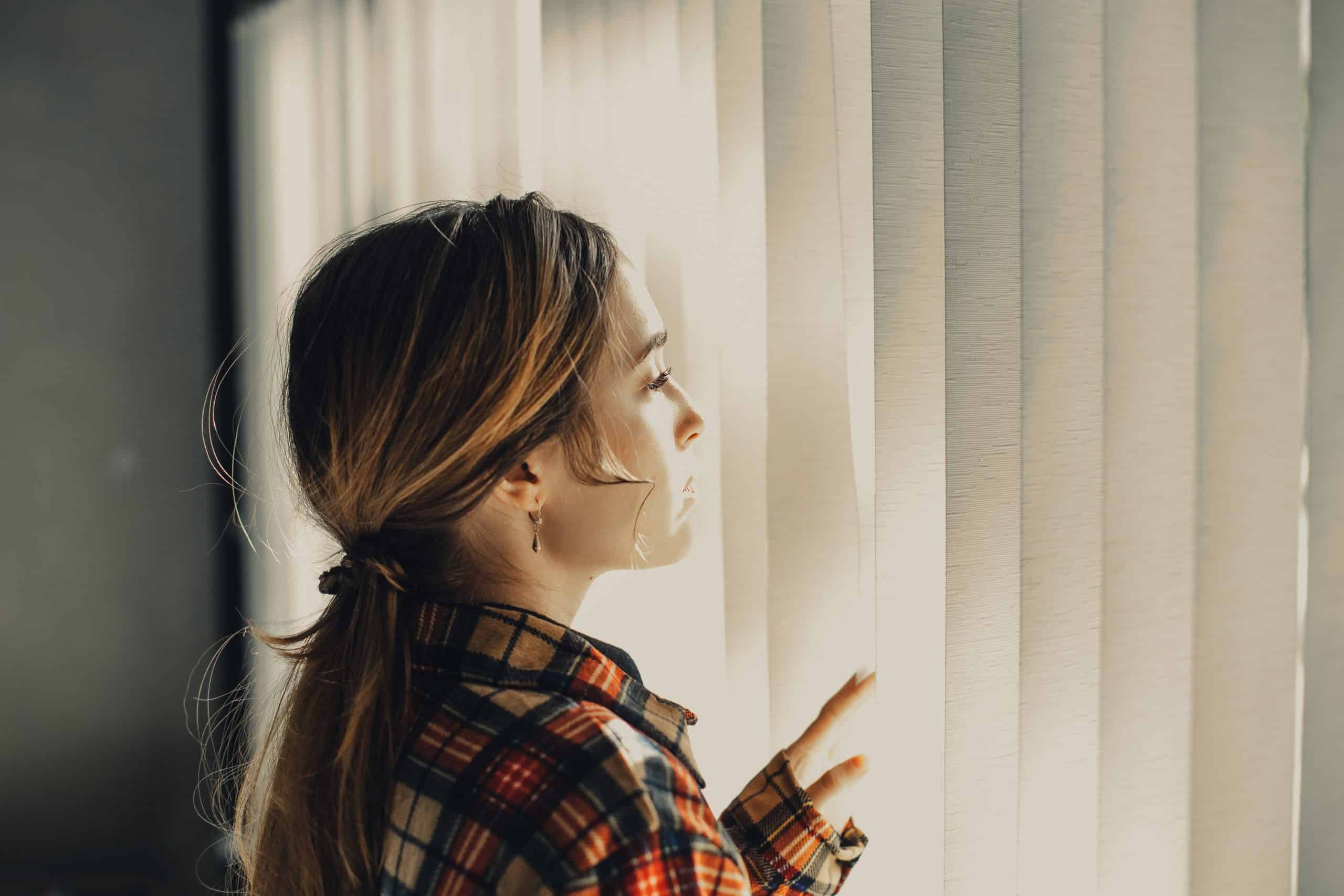 Fundersam ung kvinna som ser till och med rullgardiner av fönster i dagsljus