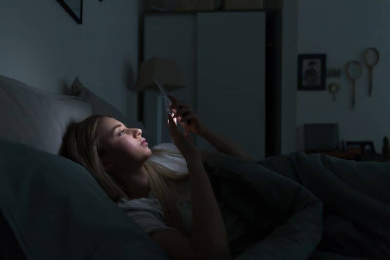 ung sömnig utmattad kvinna som ligger i sängen med hjälp av smartphonen