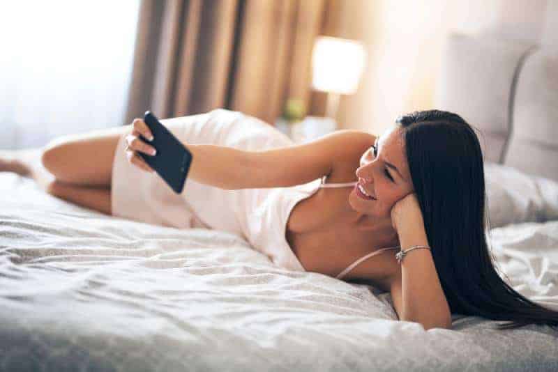 En vacker le flicka som ligger i sängen i nattklänning och håller telefonen i hennes hand