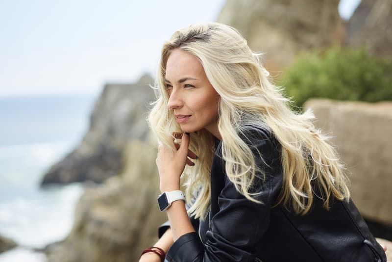 en blondin i en svart jacka står på en klippa ovanför havet och tänker