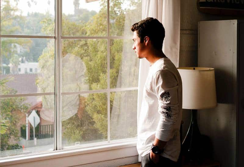 mannen i vit t-shirt står bredvid fönstret och tittar ut