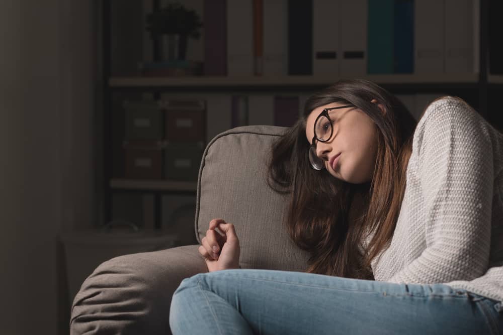 Ledsen ung kvinna med glasögon som sitter i soffan hemma, hon är deprimerad och ensam