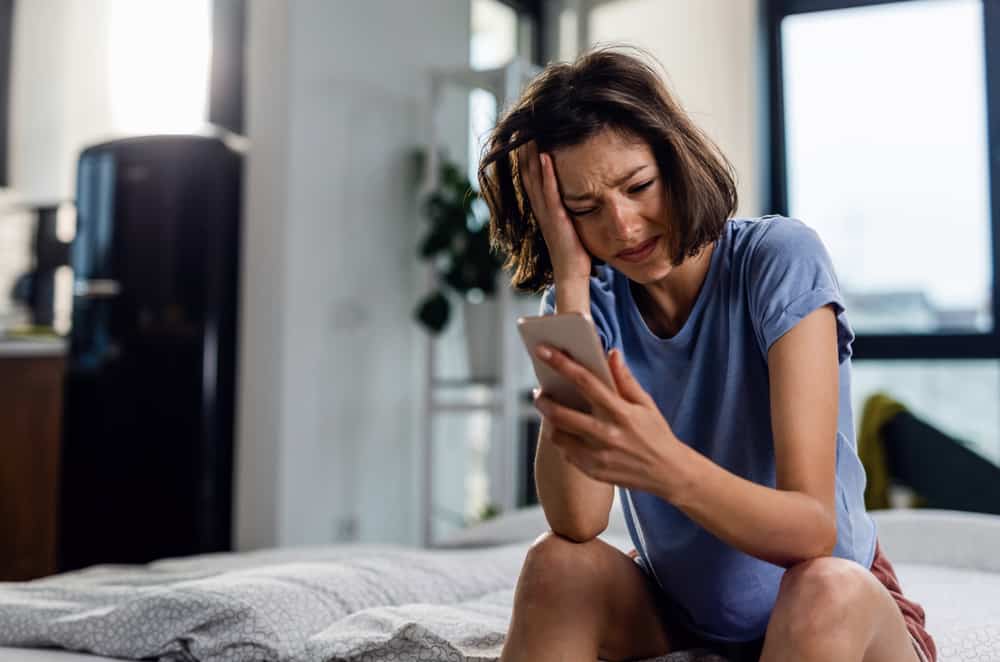 Olycklig kvinna som gråter när man sitter på sängen och läser textmeddelande