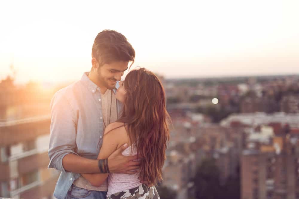 Par som är förälskade står och kramar på en byggnadstak vid solnedgången