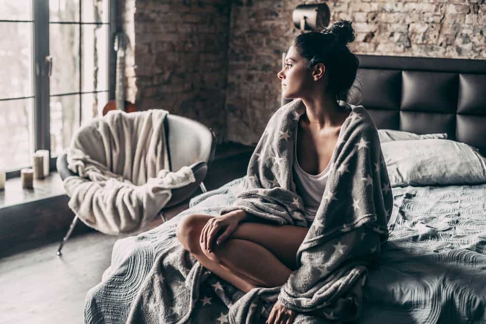 Underbar ung kvinna täckt med filt som tittar genom fönstret när man sitter i bed