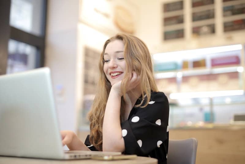 en leende kvinna i en svart polka dot T-shirt sitter vid köksbordet och använder en bärbar dator