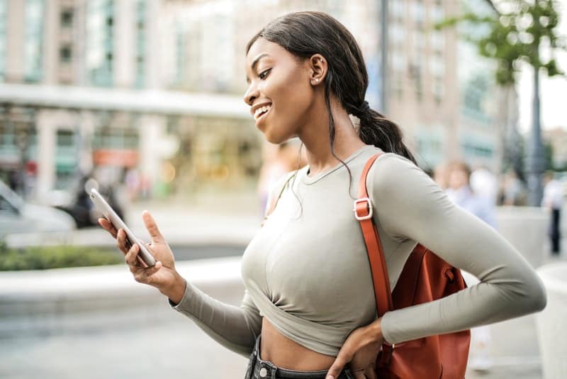 en leende svart kvinna med en brun väska på gatan använder en mobiltelefon