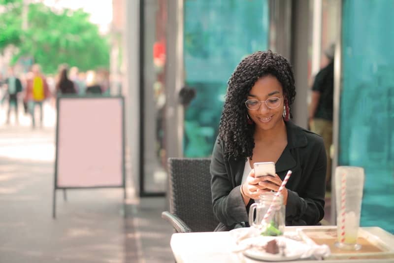 en svart kvinna sitter på terrassen på ett kafé och motsvarar via smartphone