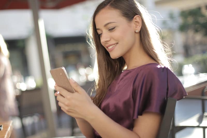 en vacker kvinna i en lila T-shirt sitter på terrassen på ett kafé och använder en mobiltelefon med ett leende