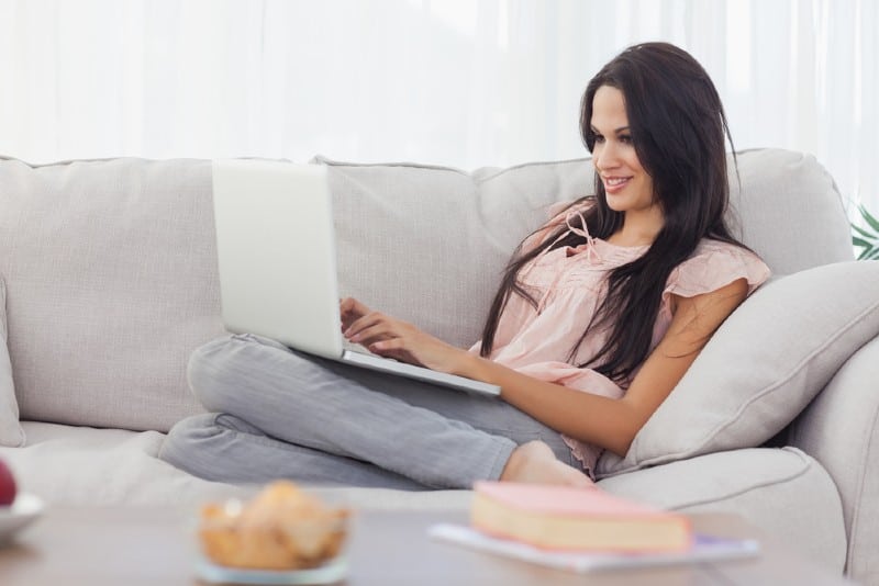 Attraktiv brunettkvinna som hemma använder hennes bärbara dator på soffan