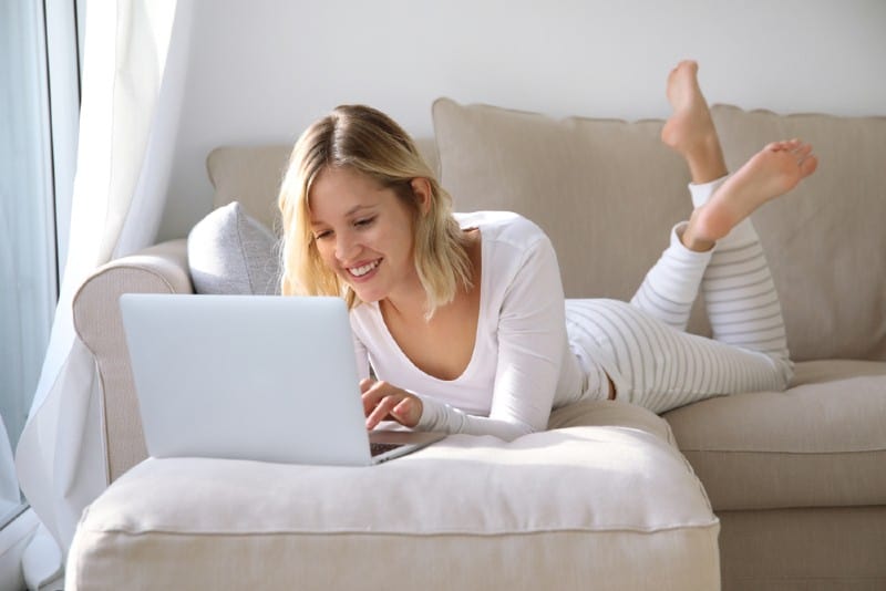 Ung kvinna som ligger på en soffa och arbetar med en bärbar dator