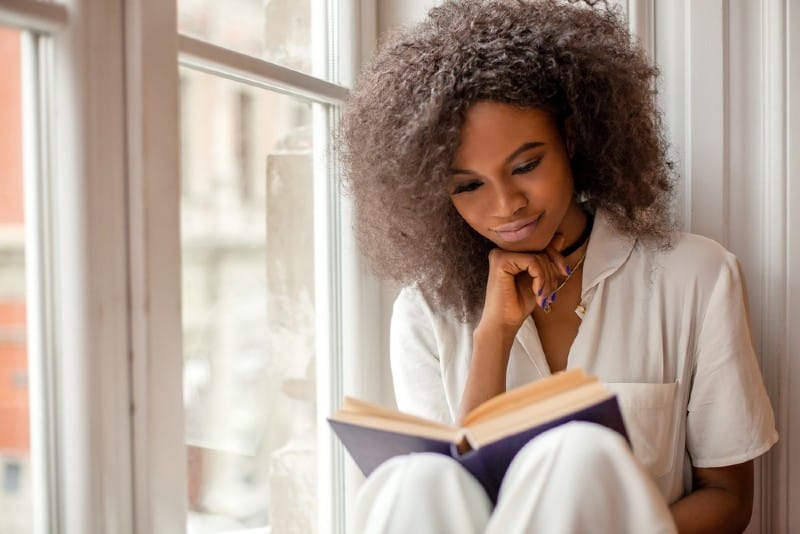 Ganska afroamerikansk flicka som läser ett bok som sitter på fönsterbrädan