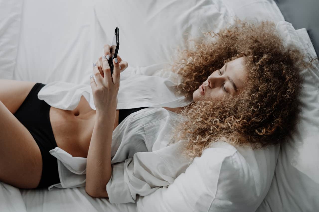 Kvinna i vit skjorta som ligger på säng och smsar