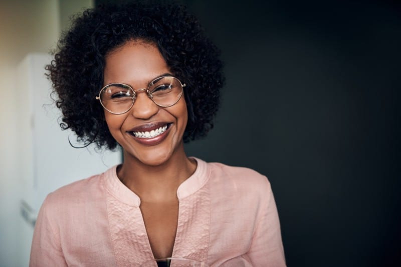 Ung afrikansk affärskvinna som bär glasögon och skrattar medan hon står ensam i ett modernt kontor