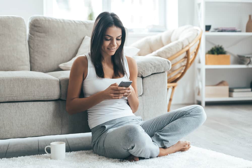 ung kvinna tittar på sin smartphone och ler medan hon sitter på mattan