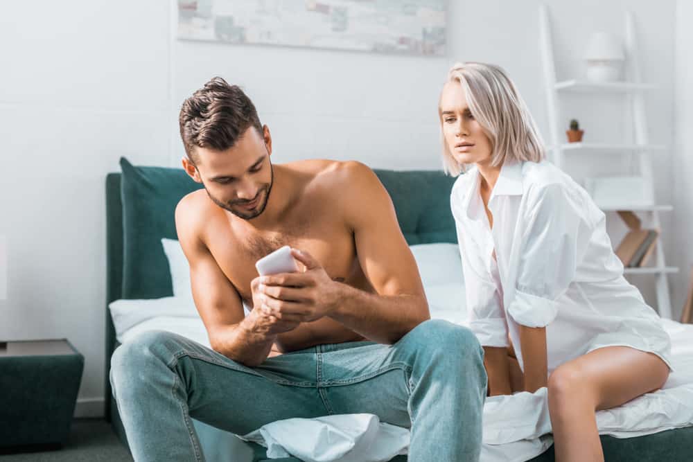 ung man använder smartphone av sin flickvän medan hon sitter på bakgrunden i sovrummet