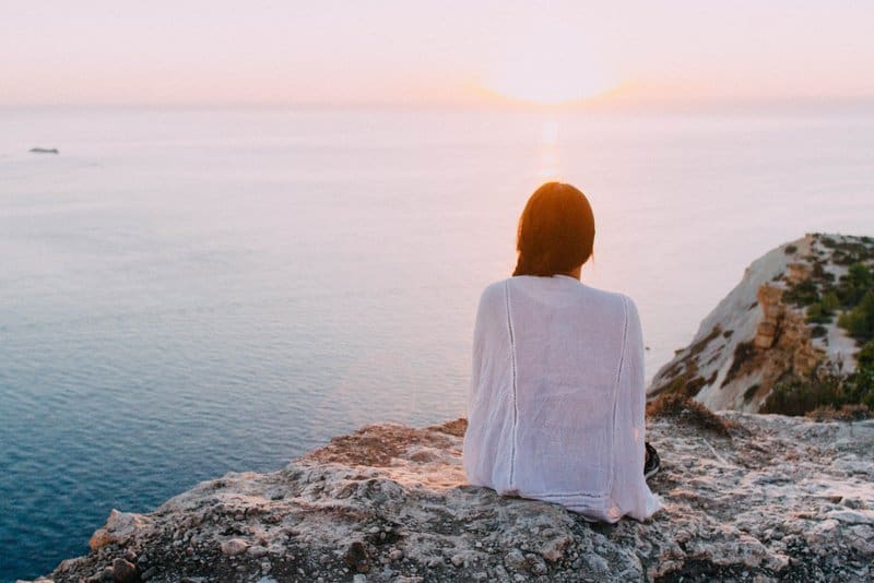 en kvinna sitter på en kulle ovanför havet och tittar på solnedgången