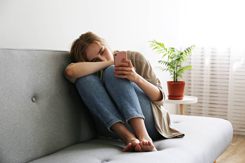 en lockigt ledsen blondin sitter i soffan och tittar på sin mobiltelefon