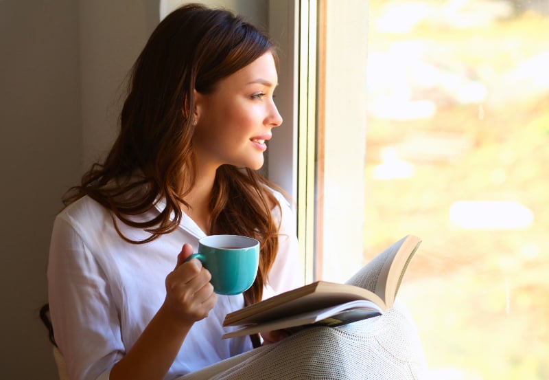 en ung kvinna som sitter vid fönstret med en bok i handen