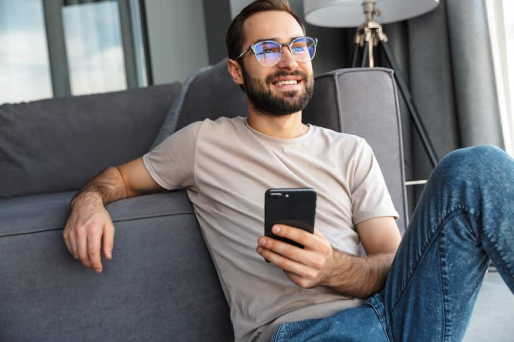 Attraktivt smart sammanträde för ung man på ett golv i vardagsrummet som använder mobiltelefonen