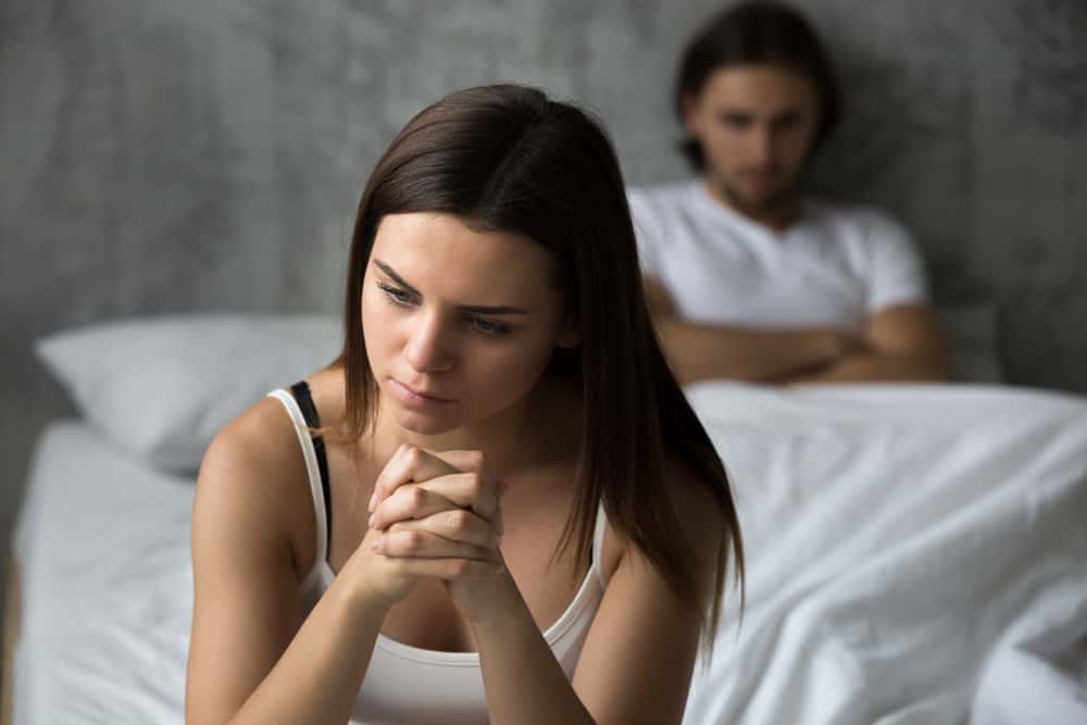 Den frustrerade ledsna flickvännen sitter på säng med mannen i bakgrunden
