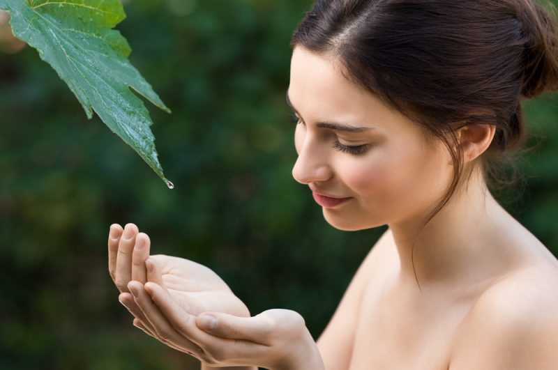 Den härliga unga kvinnan tar en droppe av klart vatten från ett blad i naturen