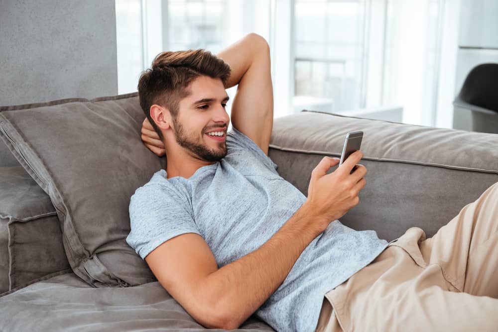 Fotoet av den lyckliga unga mannen ligger på soffan och ser på telefonen