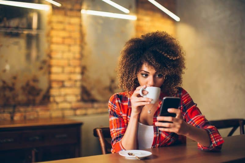 Ung kvinna på kaféet som dricker kaffe och använder mobiltelefonen