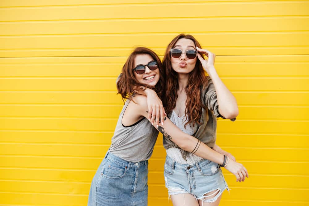 mage av två unga glada kvinnavänner som står över gul vägg