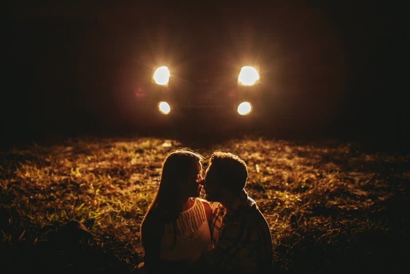 på kvällen sitter ett ungt par i ett fält framför en bil med lampor på och kyssar