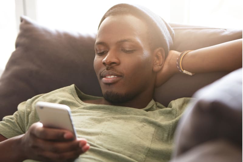 den svarta mannen ligger på soffan och använder sin smartphone med ett leende