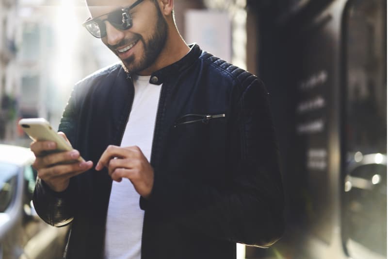 en leende man i svart jacka står på trottoaren och läser ett sms på sin smartphone