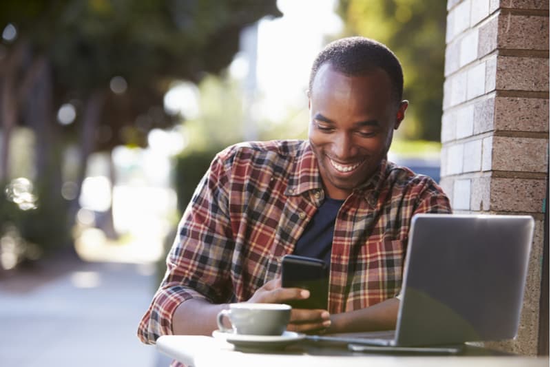 en leende svart man sitter utanför vid ett bord och använder en mobiltelefon