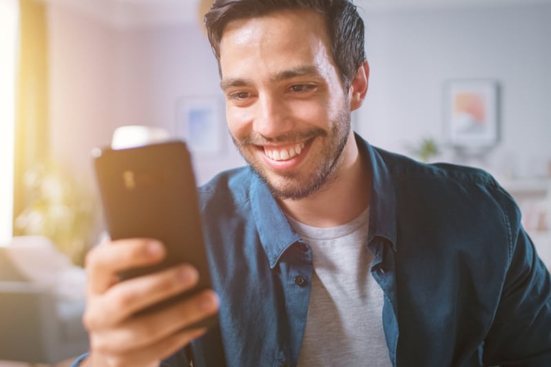 en överlycklig man som sitter i huset med ett leende som läser något på sin smartphone