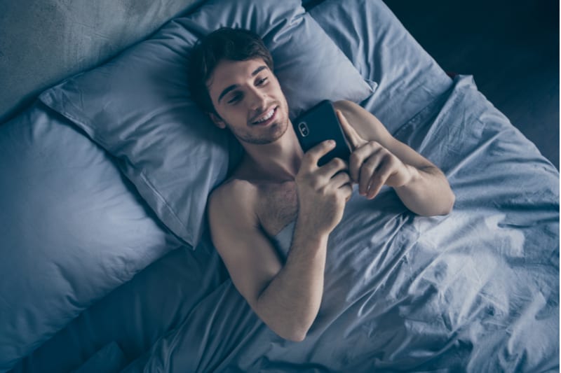 en stilig man ligger i sängen och använder en smartphone
