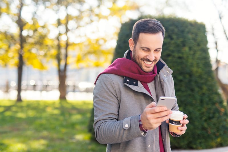 en stilig skäggig man i en grå kappa står utanför och använder en smartphone med ett leende