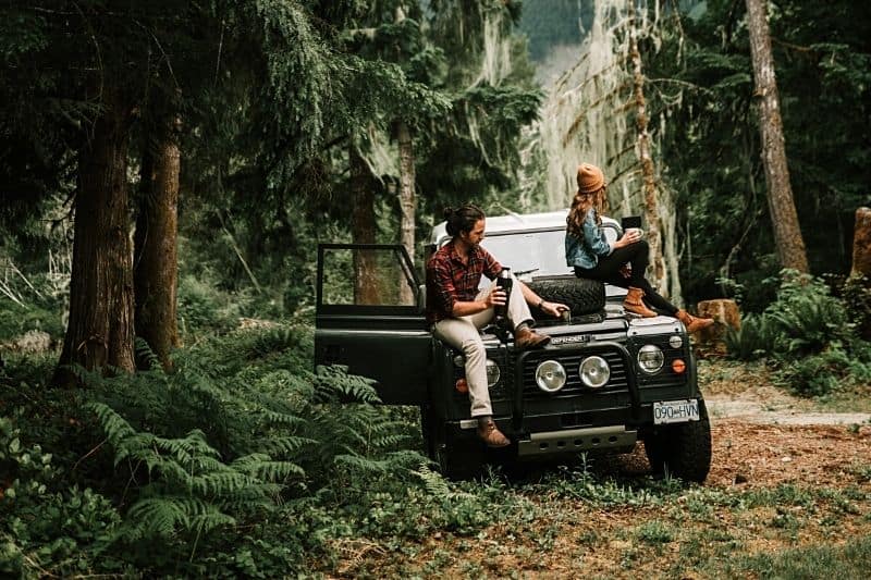 ett kärleksfullt par som sitter på huven på en jeep i skogen och dricker kaffe