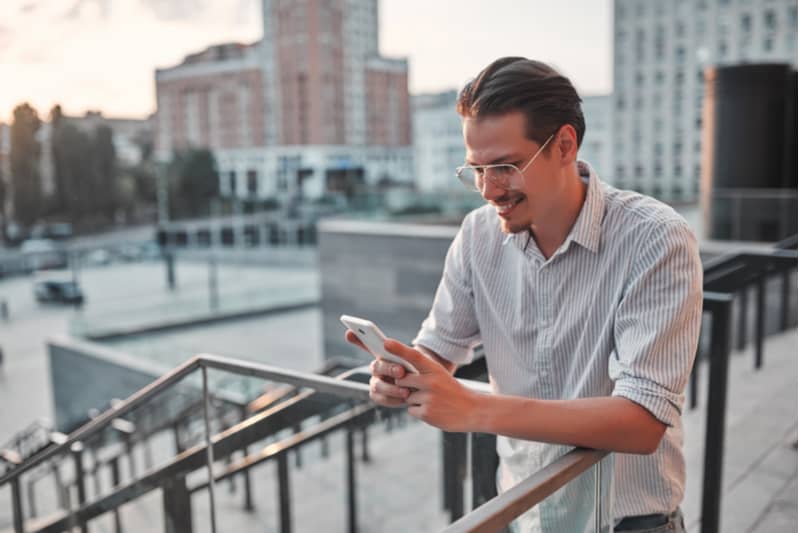 ute på terrassen står en leende man med glasögon och använder en smartphone