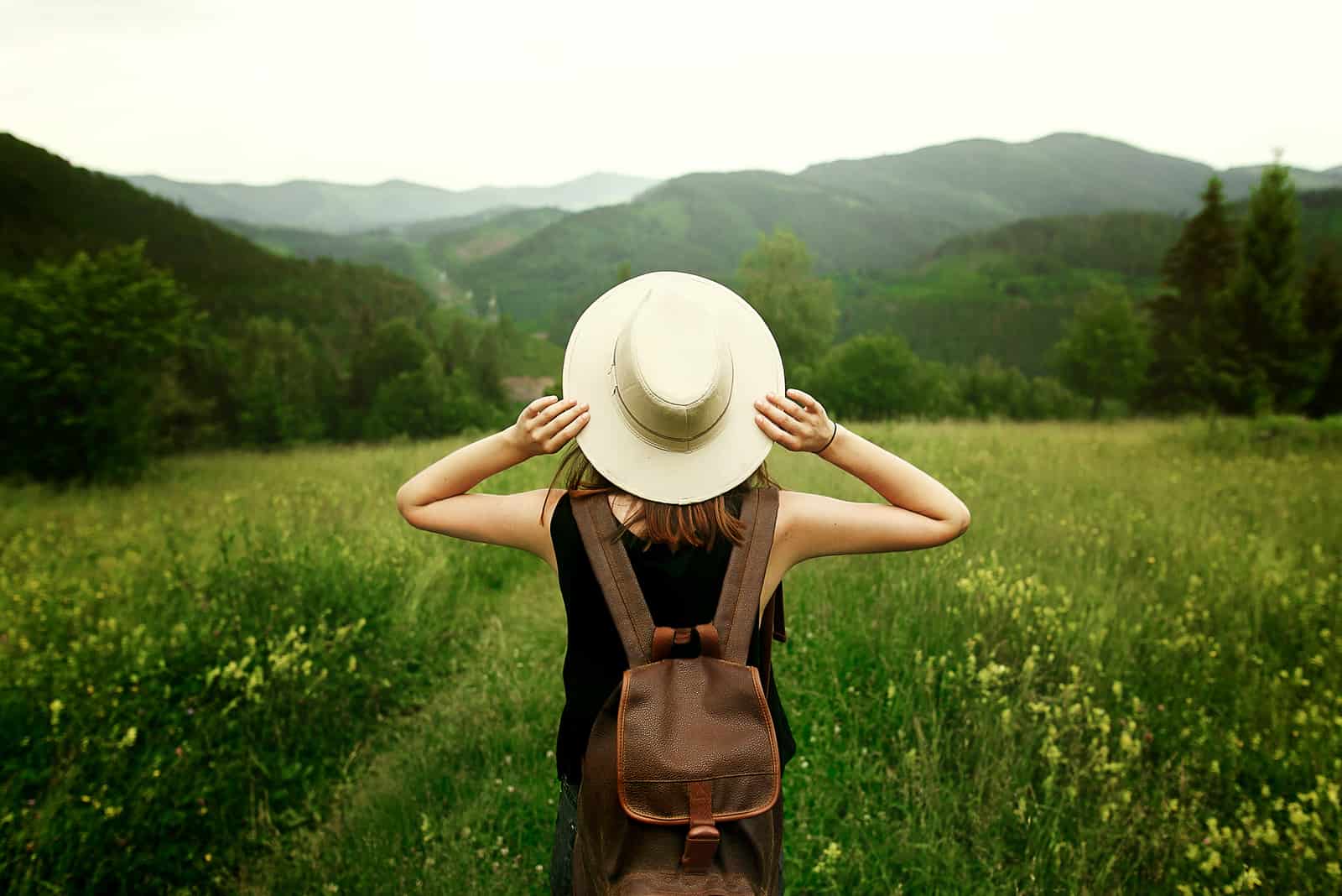 kvinna resenär med ryggsäck håller hatt och tittar på fantastiska berg och skog