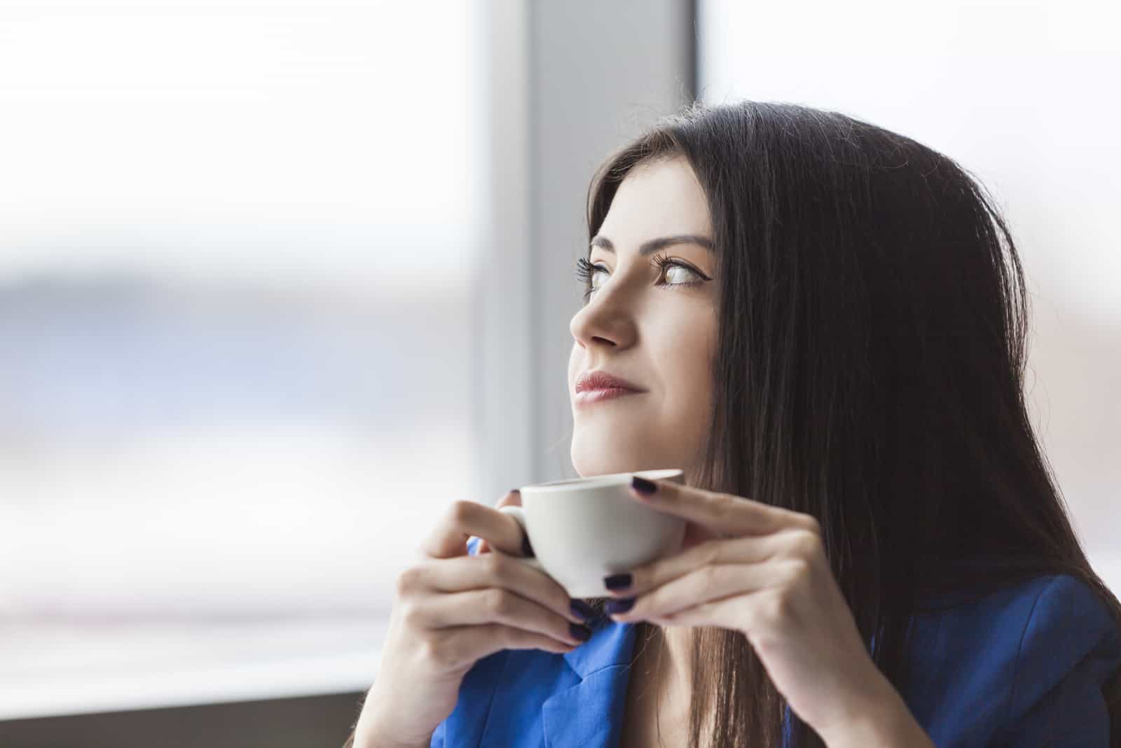 kvinna som håller en vit kaffekopp och tittar i kontorsfönstret (1)