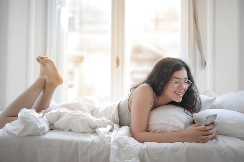 glad kvinna som ser till sin telefon medan du är i sängen