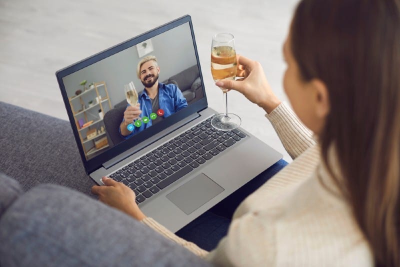 Man och kvinna med exponeringsglas vin på ett datum genom att använda bärbar datorvideochatt som sitter hemma