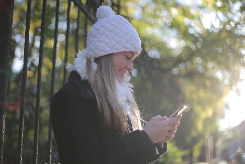 en blondin i en vintervit hatt står bredvid ett staket och skriver en sms