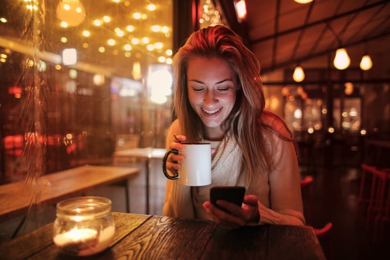en leende kvinna med en kopp te och en mobiltelefon sitter på ett café