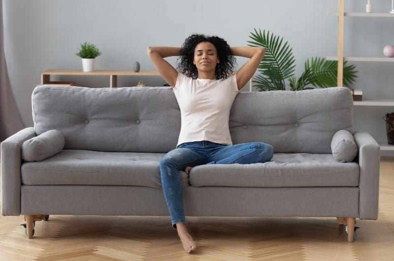 Den unga lugna svarta kvinnan som kopplar av, sitter på den bekväma soffan i modernt vardagsrum