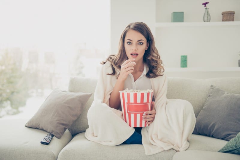 ung kvinna med pop majs i händer tittar på intressanta serier har tröst sitter på hennes säng