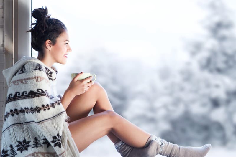 Ung vacker lycklig brunettkvinna som dricker en kopp kaffe som bär en stickad nordisk tryckponcho som sitter hemma vid fönstret