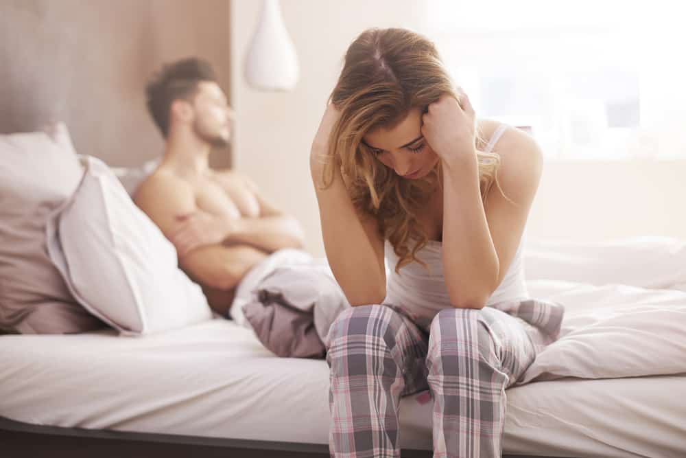 besviken kvinna som sitter på sängen med mannen i bakgrunden