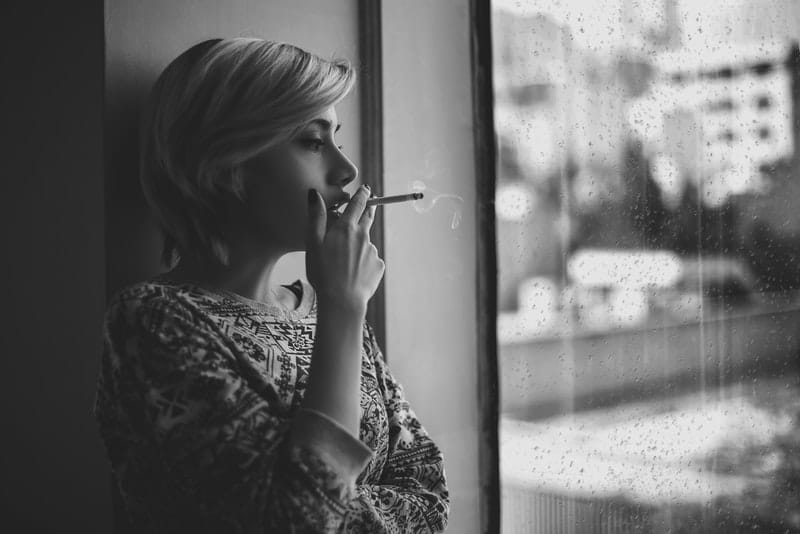 en sorglig blondin vid fönstret röker en cigarett
