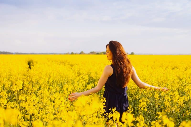 Ung kvinna i ärmlös klänning som tycker om solljus och natur på gult blommande rapsfält